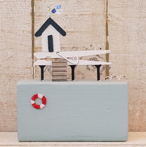 Miniature wooden beach hut model with stairs, a lifebuoy, and a patterned flag on top against a rustic wooden background.