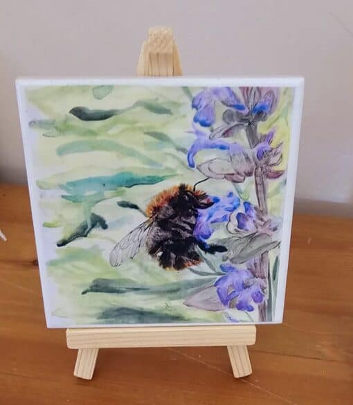 A watercolor painting of a bumblebee on a purple flower, displayed on a small wooden easel against a light background.