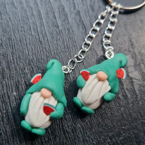 Two gnome keychains with green hats and beards, holding watermelon slices, lying on a dark textured surface.