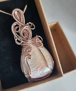 A copper wire-wrapped teardrop pendant with an intricate design, displayed in a black velvet box.