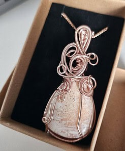 A handcrafted copper wire-wrapped pendant with an embedded polished stone, presented in a beige box with a black lining.