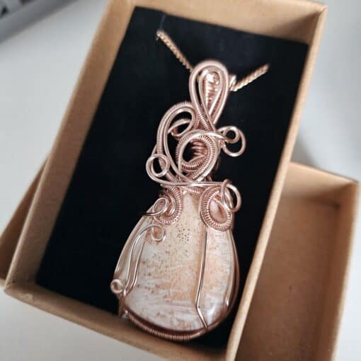 A handcrafted copper wire-wrapped pendant with an embedded polished stone, presented in a beige box with a black lining.