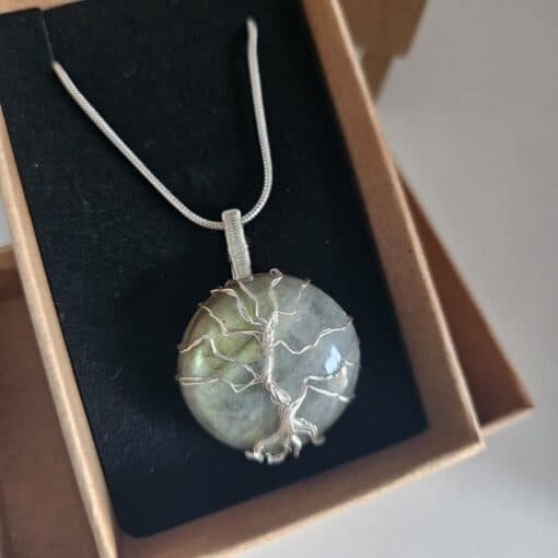 Silver pendant with a wire-wrapped tree design, set in a round labradorite stone, displayed in an open cardboard box.