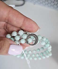 Hand holding a light green beaded bracelet with a silver circular clasp, on a white surface.