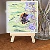 Small wooden easel displaying a painting of a bumblebee on purple flowers, with a blurred background of a glass vase and flowers.