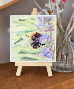 Small wooden easel displaying a painting of a bumblebee on purple flowers, with a blurred background of a glass vase and flowers.