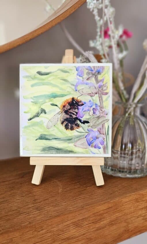 Small wooden easel displaying a painting of a bumblebee on purple flowers, with a blurred background of a glass vase and flowers.