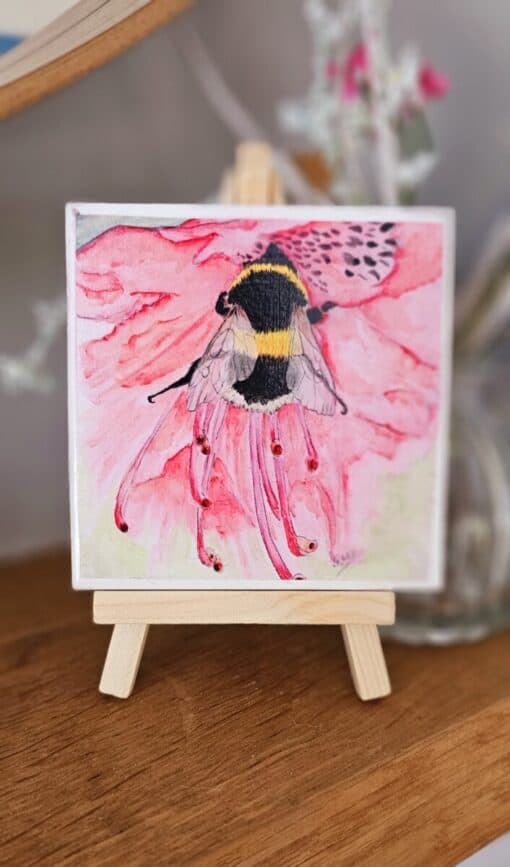 A watercolor painting of a bee on a pink flower is displayed on a small wooden easel.