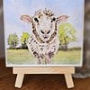 A small easel displays a painting of a sheep standing on green grass with trees and a blue sky in the background.