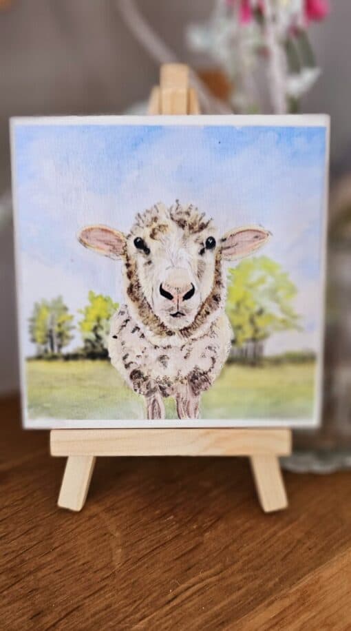 A small easel displays a painting of a sheep standing on green grass with trees and a blue sky in the background.