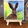 Watercolor painting of a rabbit on a small easel, with a colorful background and floral accents.