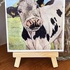 Painting of a cow with black and white spots on a small wooden easel, displayed on a wooden surface with flowers in the background.