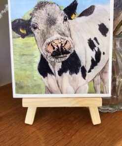 Painting of a cow with black and white spots on a small wooden easel, displayed on a wooden surface with flowers in the background.