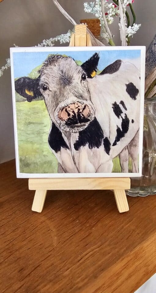 Painting of a cow with black and white spots on a small wooden easel, displayed on a wooden surface with flowers in the background.