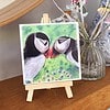 Painting of two puffins touching beaks on a small easel, displayed on a wooden shelf next to a vase with flowers.