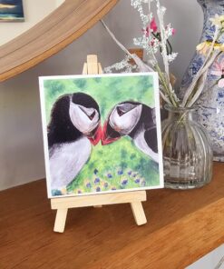 Painting of two puffins touching beaks on a small easel, displayed on a wooden shelf next to a vase with flowers.