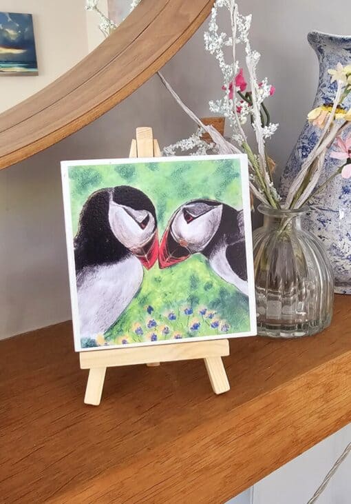 Painting of two puffins touching beaks on a small easel, displayed on a wooden shelf next to a vase with flowers.