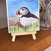 A colorful puffin drawing on a small easel, placed on a wooden shelf with a vase of white and pink flowers in the background.