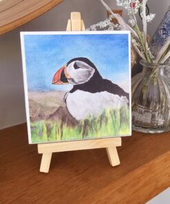 A colorful puffin drawing on a small easel, placed on a wooden shelf with a vase of white and pink flowers in the background.