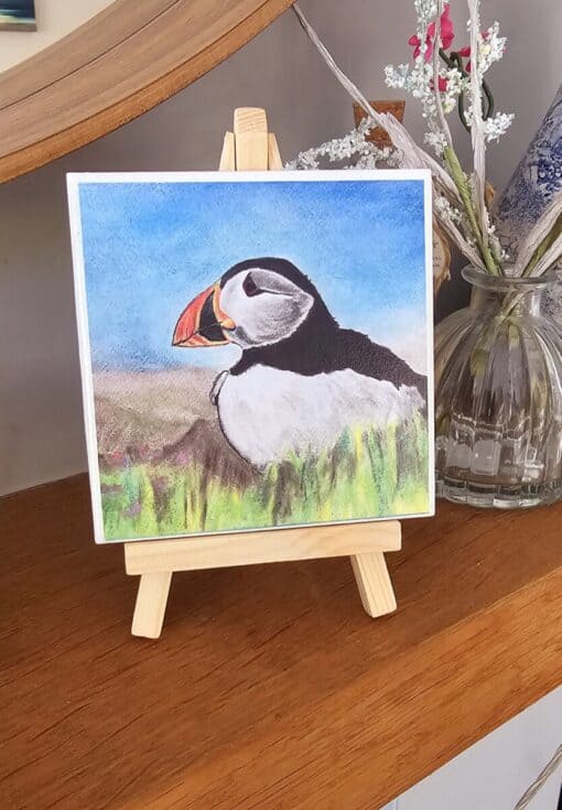 A colorful puffin drawing on a small easel, placed on a wooden shelf with a vase of white and pink flowers in the background.