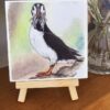 A watercolor painting of a puffin on a small easel, placed on a wooden surface with flowers in a vase behind it.