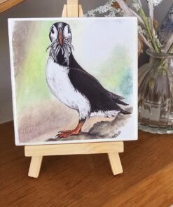 A watercolor painting of a puffin on a small easel, placed on a wooden surface with flowers in a vase behind it.