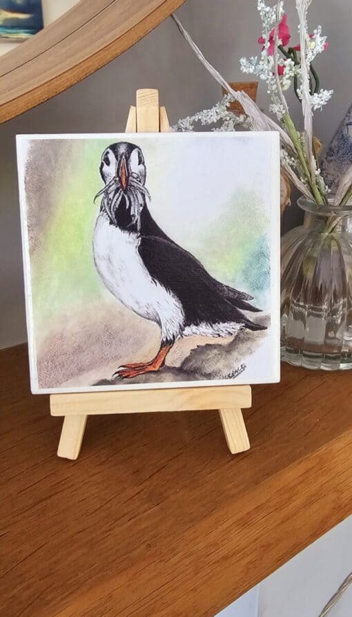 A watercolor painting of a puffin on a small easel, placed on a wooden surface with flowers in a vase behind it.