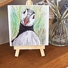 A hand-drawn puffin illustration on a mini wooden easel, placed on a shelf next to a glass vase with flowers.