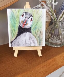 A hand-drawn puffin illustration on a mini wooden easel, placed on a shelf next to a glass vase with flowers.