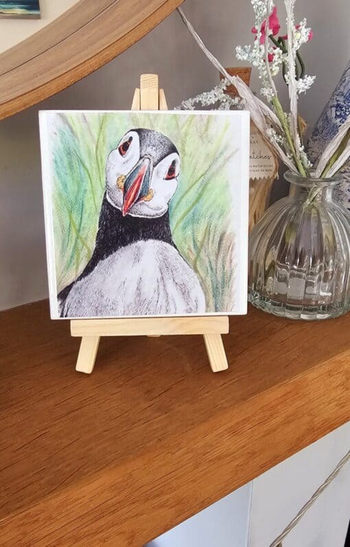 A hand-drawn puffin illustration on a mini wooden easel, placed on a shelf next to a glass vase with flowers.