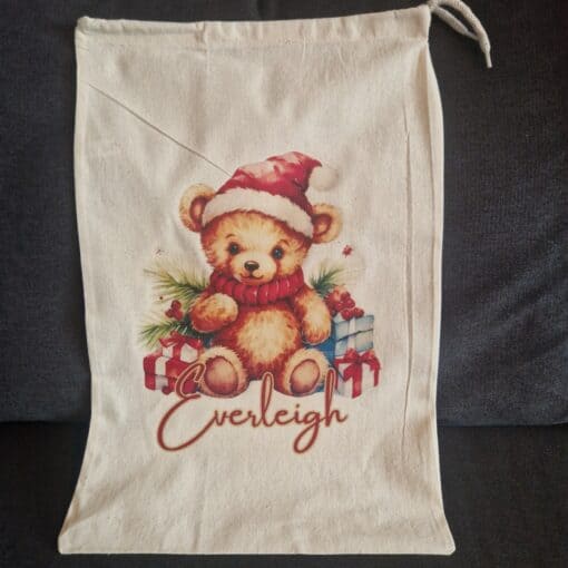 Personalised drawstring Christmas backpack with a cute teddy bear in a Santa hat, surrounded by gifts, and the name "Everleigh.