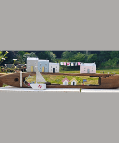 A wooden boat model holding small house figurines and a miniature clothesline, set outdoors with greenery in the background.