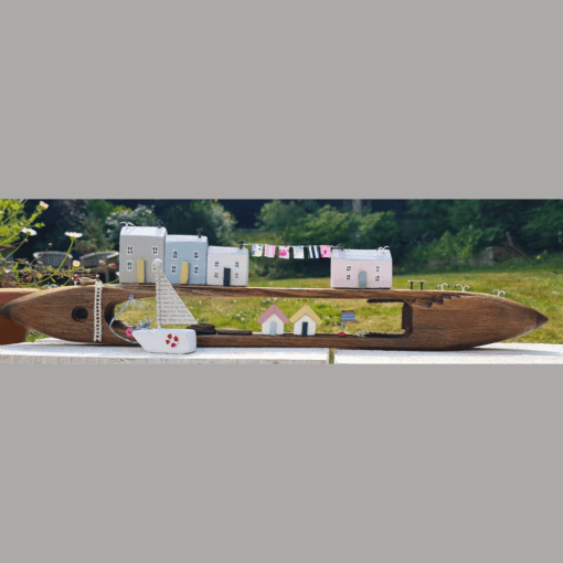 A wooden boat model holding small house figurines and a miniature clothesline, set outdoors with greenery in the background.