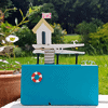 Handmade miniature house model with a pink flag on a vibrant blue base, set outdoors with greenery in the background.