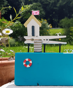 Handmade miniature house model with a pink flag on a vibrant blue base, set outdoors with greenery in the background.