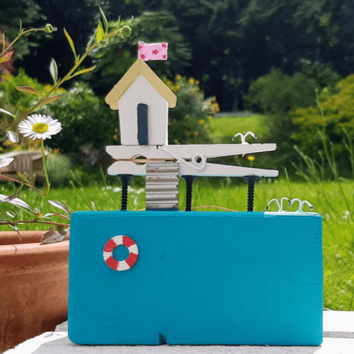 Handmade miniature house model with a pink flag on a vibrant blue base, set outdoors with greenery in the background.