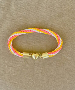 Colorful braided bracelet with pink, yellow, and white strands, and a gold heart clasp, lying on a beige surface.