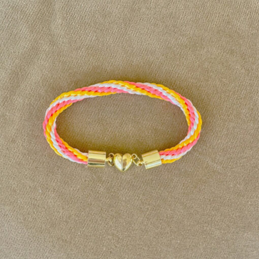 Colorful braided bracelet with pink, yellow, and white strands, and a gold heart clasp, lying on a beige surface.