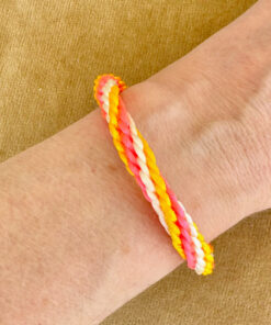 Person wearing braided bracelet with orange, pink, white, and yellow threads on their wrist, against a beige background.