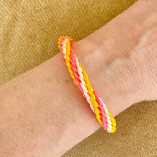 Person wearing braided bracelet with orange, pink, white, and yellow threads on their wrist, against a beige background.