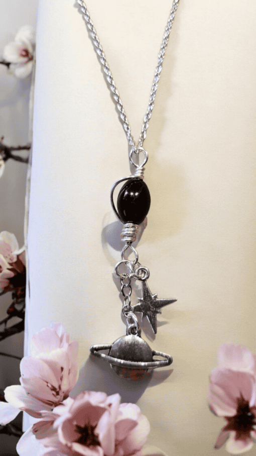 Silver necklace with black bead and charms of a star and Saturn, displayed against a neutral background with pink flowers.