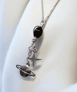 Silver necklace with black bead, and charms depicting a planet with rings and a star, displayed on a white fabric background.