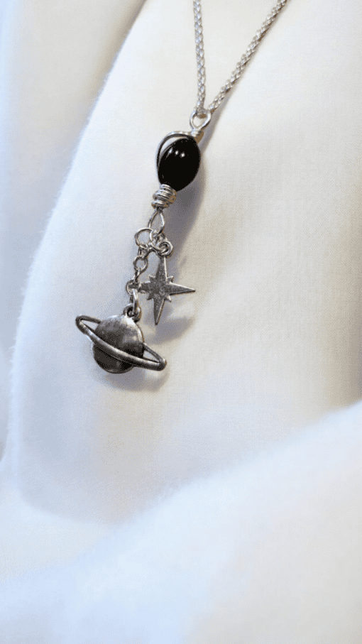 Silver necklace with black bead, and charms depicting a planet with rings and a star, displayed on a white fabric background.