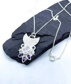 Silver pendant necklace with crescent moon design, featuring a square gemstone, star accents, and purple teardrop gems on a black slate.