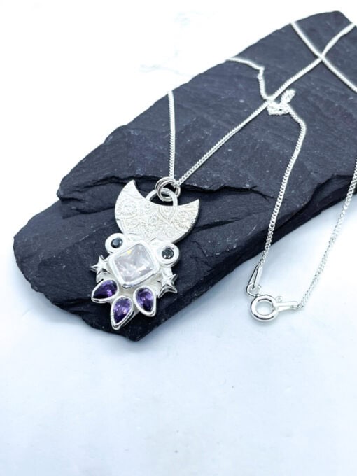 Silver pendant necklace with crescent moon design, featuring a square gemstone, star accents, and purple teardrop gems on a black slate.