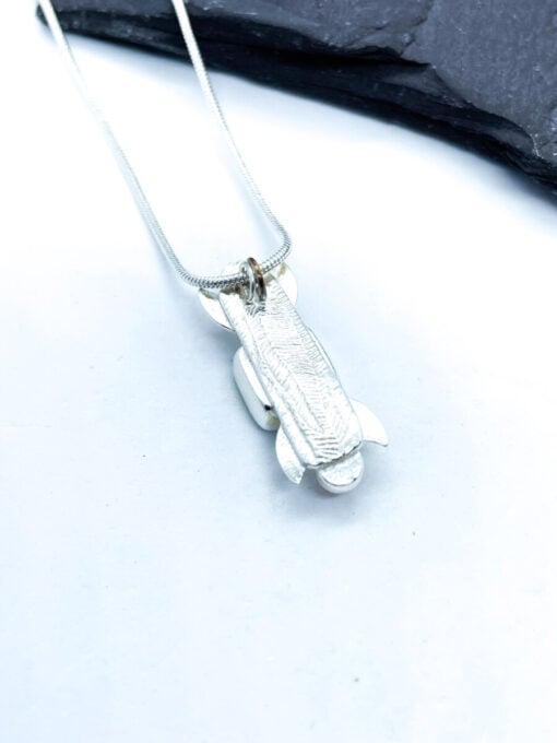 Silver pendant shaped like a rocket on a chain, placed on a white surface with a dark stone background.