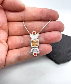 Hand holding a silver pendant necklace with intricate designs, featuring a central square gemstone and two small round orange gems.