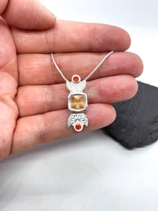 Hand holding a silver pendant necklace with intricate designs, featuring a central square gemstone and two small round orange gems.