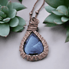 A wire-wrapped necklace with a teardrop-shaped blue gemstone pendant, placed between two green succulents.