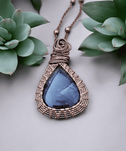 A wire-wrapped necklace with a teardrop-shaped blue gemstone pendant, placed between two green succulents.